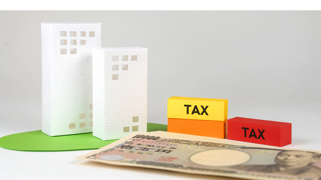 紙で出来たビルの模型と一万円札とTAXと書かれた積み木が机に置かれているイラスト